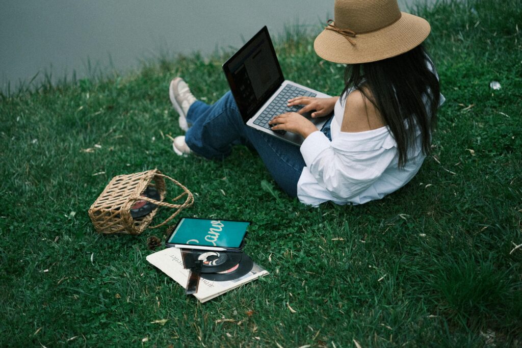 A woman sits on the grass by a lake, using a laptop. Enjoys outdoor work.